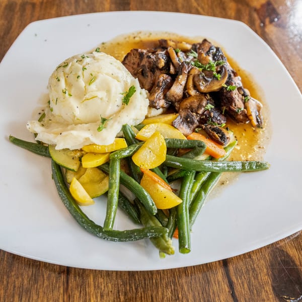 A white plate with steak and mushrooms, mashed potatoes, and mixed green and yellow vegetables.