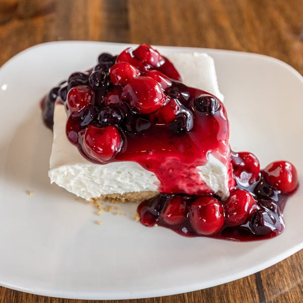 A slice of cheesecake topped with a vibrant red and purple berry compote on a white plate.