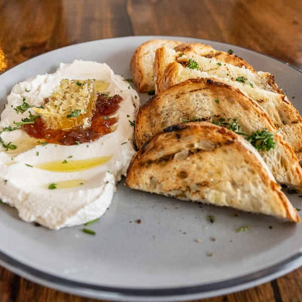 Whipped ricotta with honeycomb, fig jam, olive oil, and grilled bread on a gray plate.